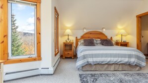 A bedroom with a large window, a bed with a striped blanket, two bedside tables with lamps, and a rug on the floor. - Ski In Ski Out in style on Village Chair (Jay)