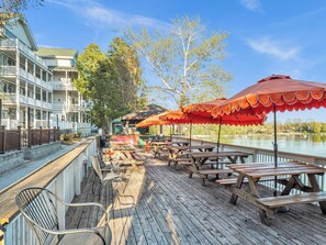 A wooden deck with picnic tables and chairs, overlooking a body of water, with a building featuring balconies in the background.