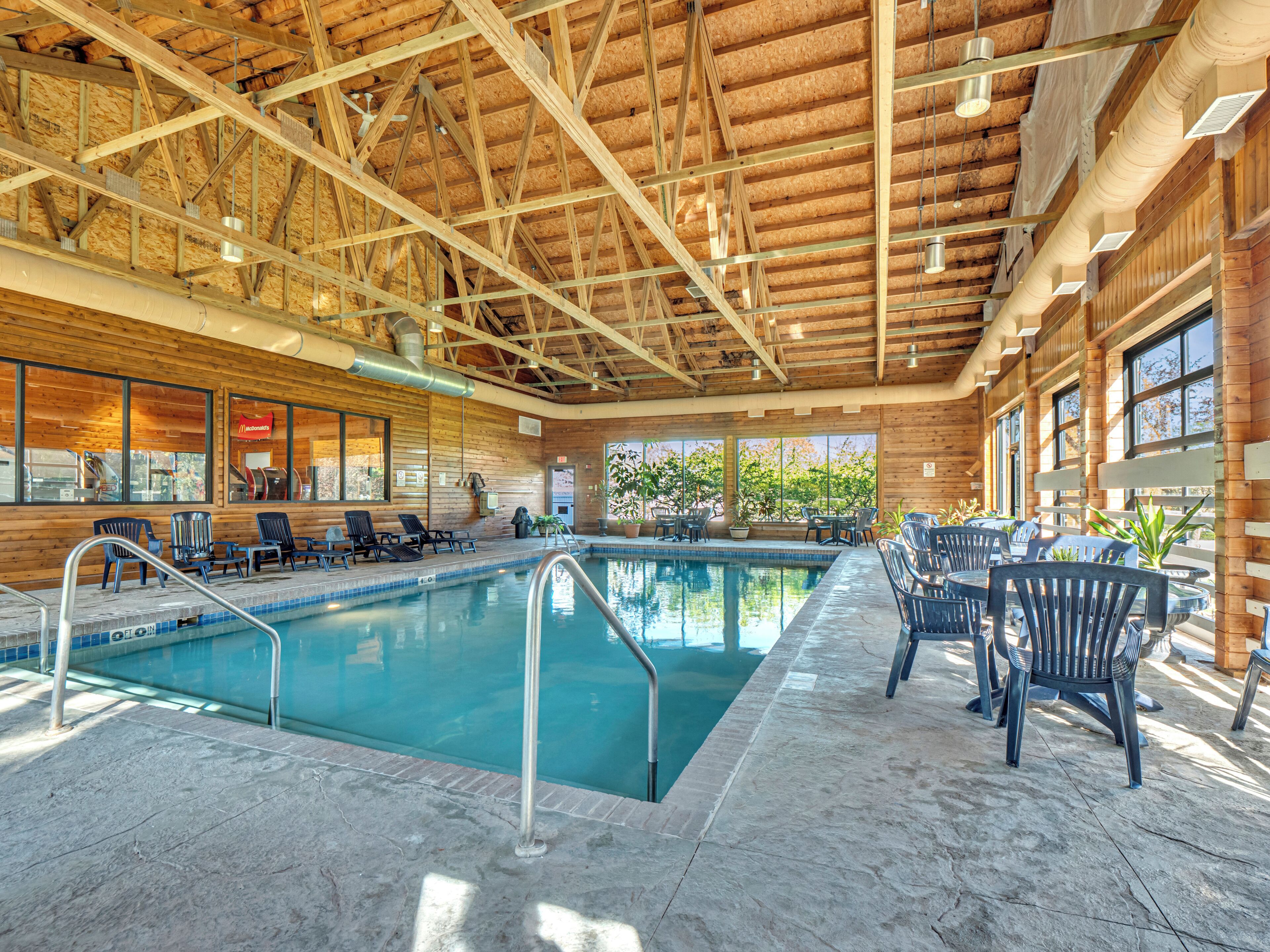 An indoor swimming pool with a wooden ceiling and walls, surrounded by lounge chairs and tables.