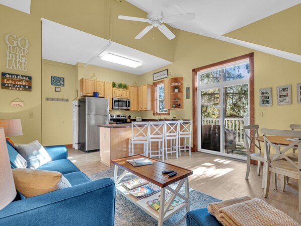 A spacious living room with a blue sofa, a wooden coffee table, and a kitchen area with wooden cabinets and a refrigerator.
