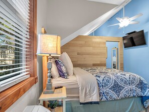A bedroom with a bed, a nightstand, a lamp, a TV, and a window with blinds.