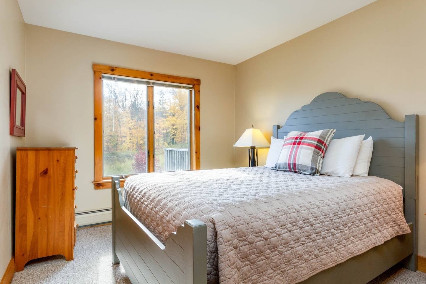 A bedroom with a bed, a wooden nightstand, a lamp, and a window with a view of trees.