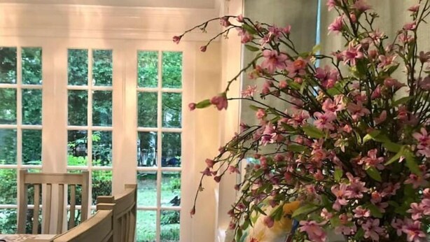 Ein Zimmer mit einem Holzesstisch, einem Stuhl, einer Vase mit rosa Blumen und einem Fenster mit Blick auf Grünflächen.