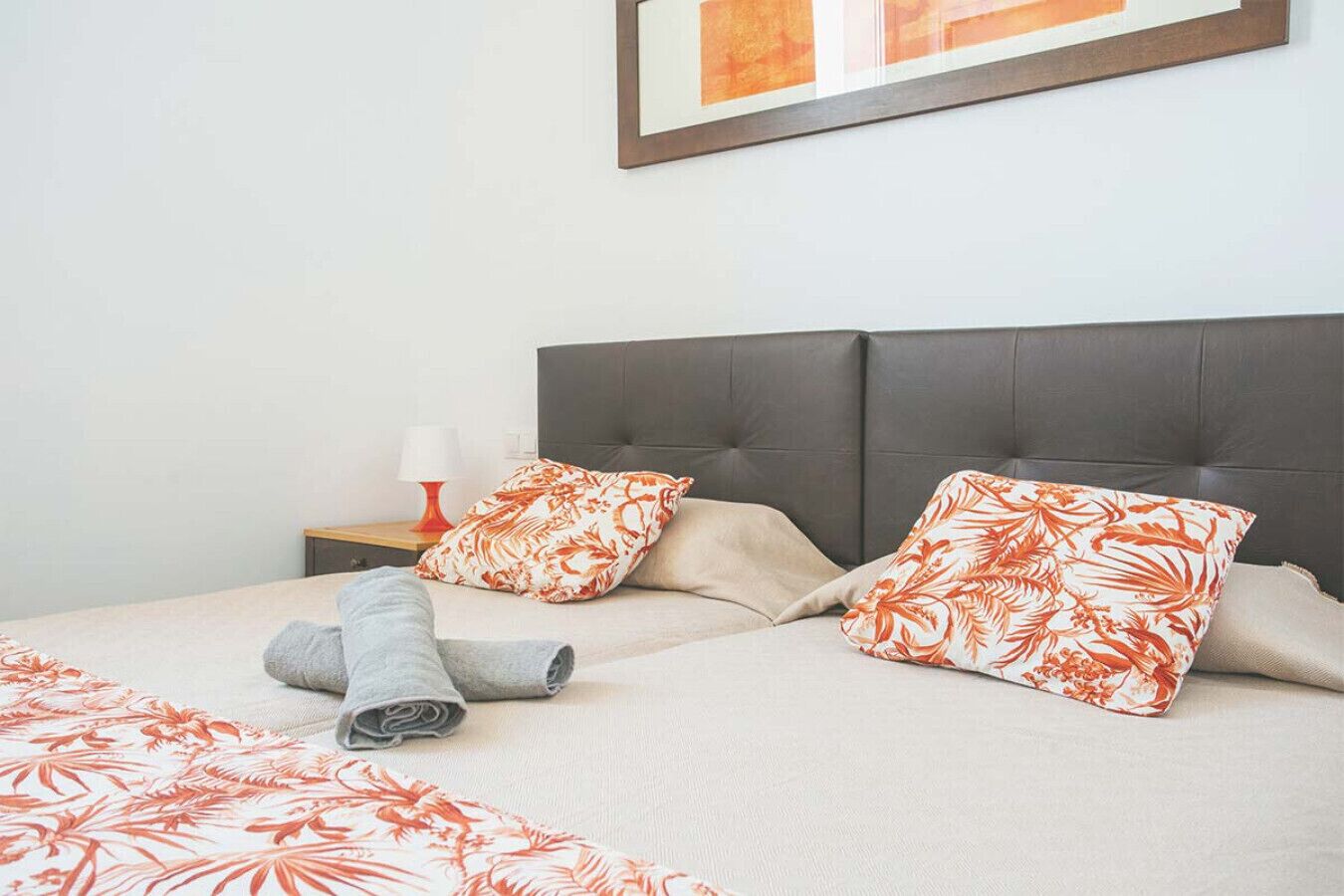 Ein ordentlich bezogenes Bett mit blumigem Kissenmuster und einem gefalteten Handtuch, ein Nachttisch mit Lampe und ein gerahmtes Bild an der Wand.