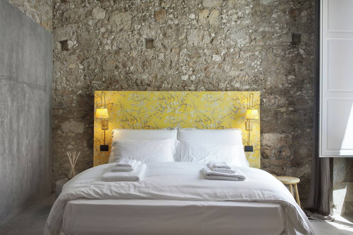 A bed with white linens and towels, a stone wall, and a yellow floral headboard.