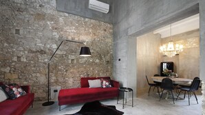 A modern living room with a stone wall, red sofa, black floor lamp, and a dining area with a chandelier. - Isonzo Suites (Syracuse)