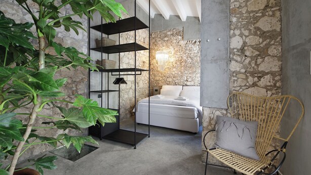 A bedroom with a stone wall, a bed, a black shelving unit, a wicker chair, and a large green plant. - Isonzo Suites (Syracuse)