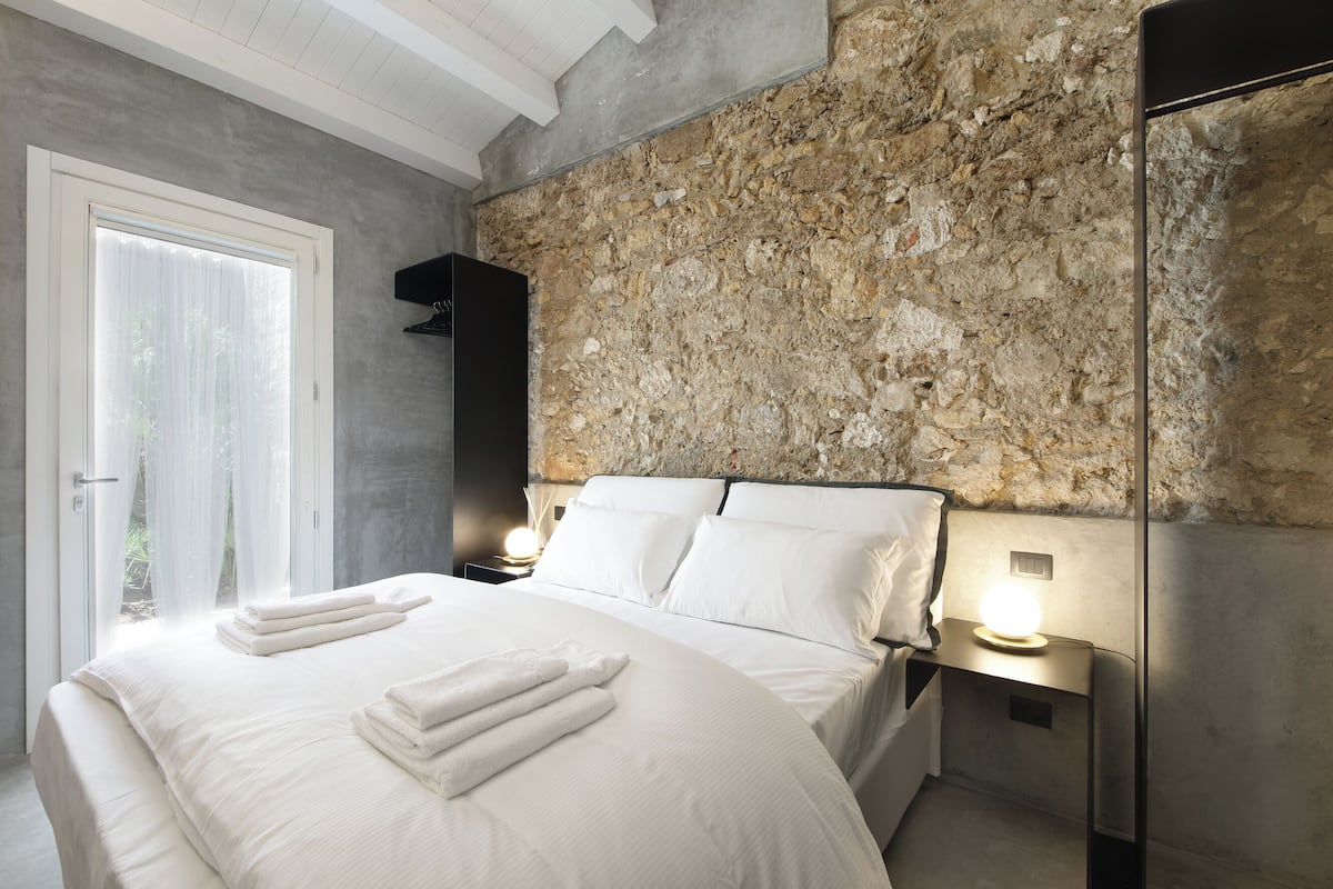 A bedroom with a stone wall, a bed with white linens, two bedside lamps, and a door leading to an outdoor area.
