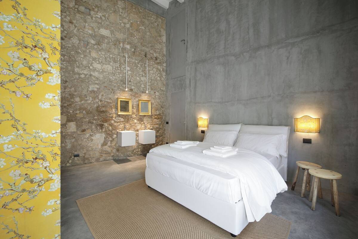 A bedroom with a stone wall, a bed with white linens, two bedside tables, and wall-mounted lights.