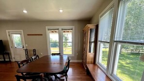 Un espace repas avec une grande table et des chaises en bois, un banc placé le long de la fenêtre et une vue sur une cour verdoyante.