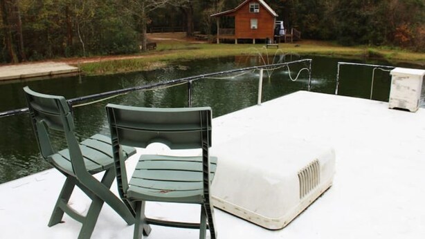 A dock with two green chairs and a white heater, overlooking a calm body of water with a wooden cabin in the background. - A Shore Bet 36' Vintage Houseboat. (Bogalusa)