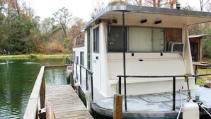 A houseboat docked at a wooden pier with a covered deck area and windows. - A Shore Bet 36' Vintage Houseboat. (Bogalusa)