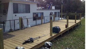 A small, single-story building with a wooden deck and a fenced area. - A Shore Bet 36' Vintage Houseboat. (Bogalusa)