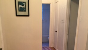 Un pasillo con suelo de madera y paredes blancas que conduce a un baño, donde se puede ver un inodoro a través de una puerta entreabierta.