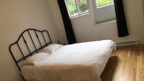 Una cama individual con estructura metálica en una habitación con suelo de madera y una ventana con cortinas negras.