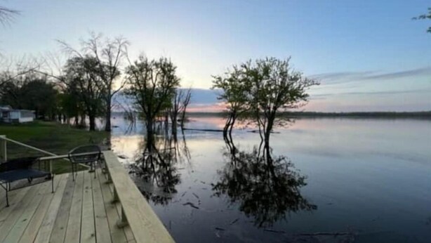 Une jetée en bois s’étendant sur une étendue d’eau calme, où se reflètent les arbres, et entourée d’une zone herbeuse.