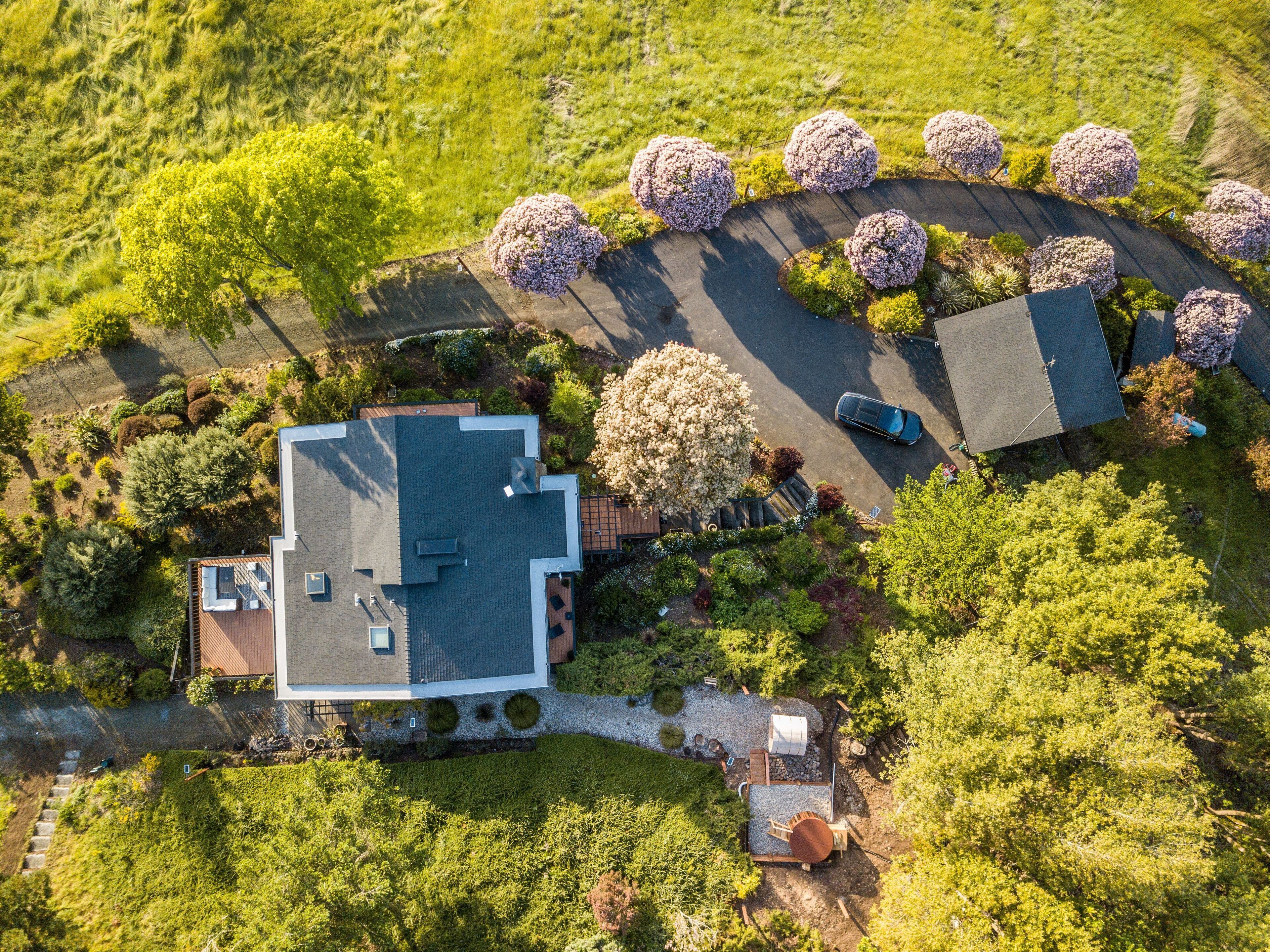 An aerial view of a modern house with a large roof, surrounded by lush greenery and flowering trees.