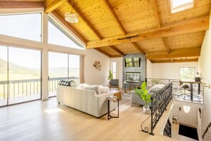 Un salón amplio con techos altos, ventanas grandes, chimenea y un balcón con vistas a las colinas.