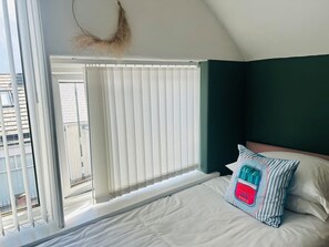 A bedroom with a bed, a window with blinds, a view of the outside, and a decorative hanging.
