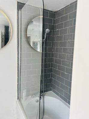 A corner bathroom with a glass shower enclosure, a white bathtub, and a round mirror.