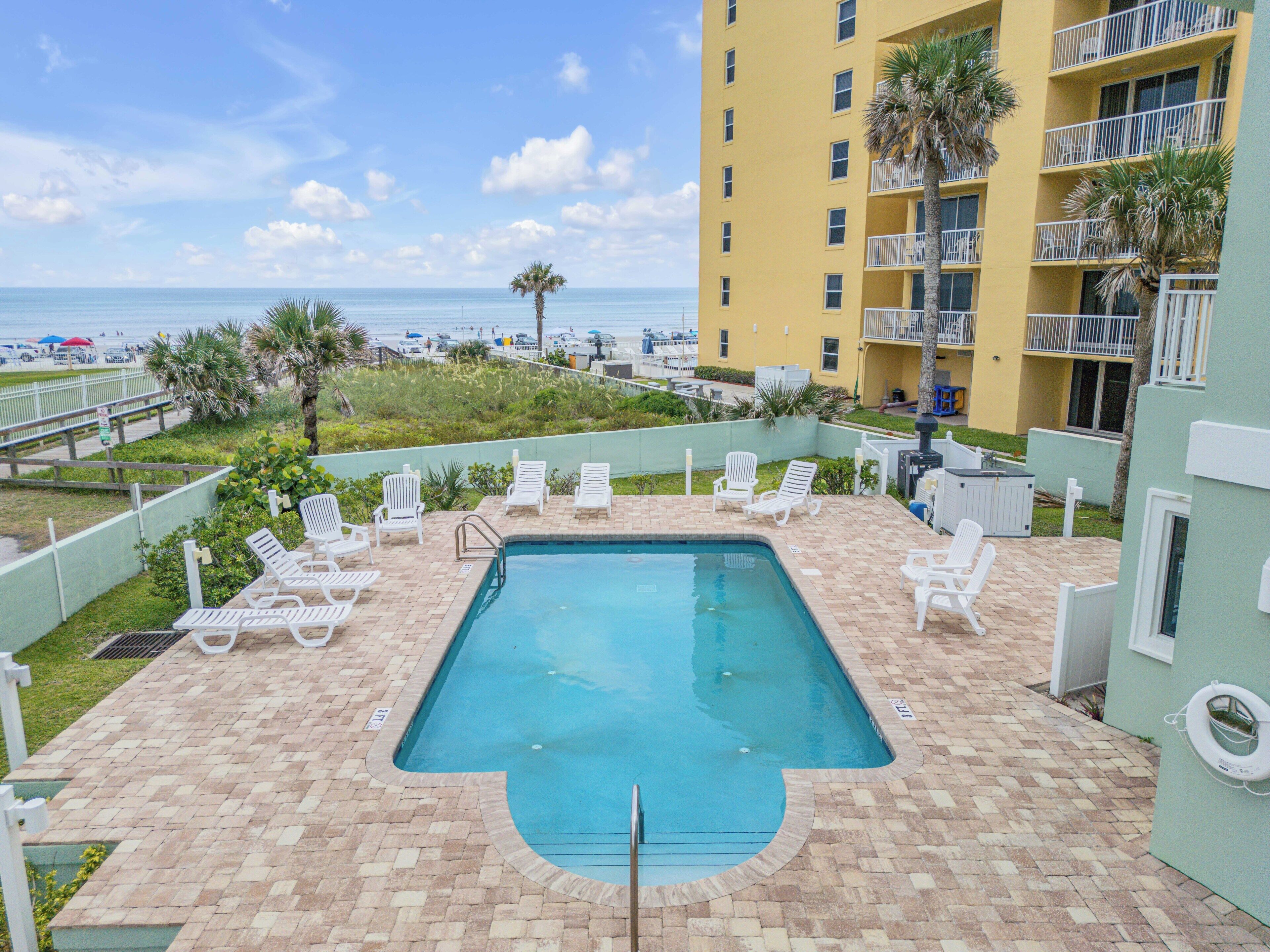 A swimming pool with lounge chairs, a view of the beach, and a building with balconies.