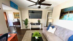 A living room with a ceiling fan, a TV mounted on the wall, a sofa with cushions, a coffee table with a plant, and a desk with a lamp. - Main St. Retreat (Safety Harbor)
