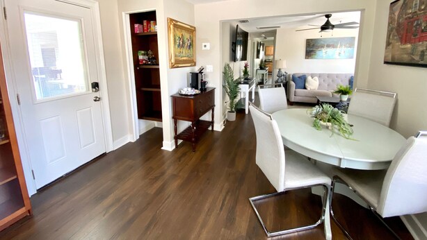 A dining area with a white table, chairs, and a potted plant. There is a wooden cabinet with a painting above it and a ceiling fan in the background. - Main St. Retreat (Safety Harbor)
