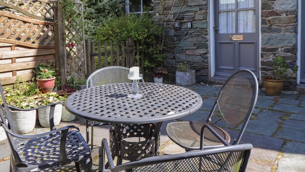 Een stenen gebouw met een houten deur en een raam. Er staat een ronde metalen tafel met stoelen en een kaars op.