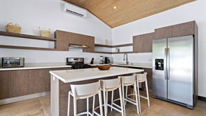 Een moderne keuken met een centraal eiland, witte barkrukken, een roestvrijstalen koelkast en houten keukenkastjes.