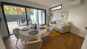 A dining area with a round table set for four, a sofa, and a large window offering a view of a garden. - Le Pied à Terre N°3 (Grosseto-Prugna)