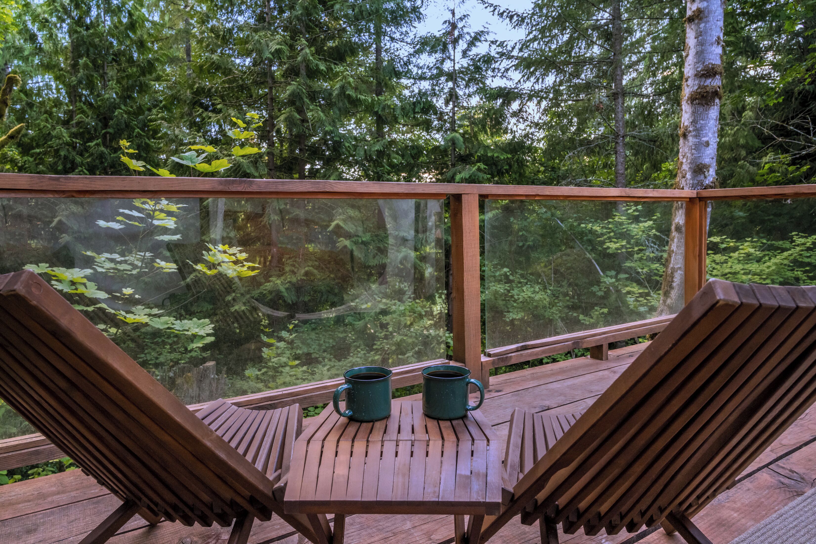 A wooden deck with two lounge chairs and two green mugs on a table, overlooking a forested area.