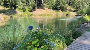 Une terrasse en bois mène à un étang paisible, entouré d’une végétation luxuriante et de fleurs bleues.