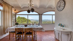 A dining area with a table set for two, a chandelier, a wall clock, and a view of the landscape through large windows. - VERDI: House with spectacular sea views and private pool in Pals (Pals)