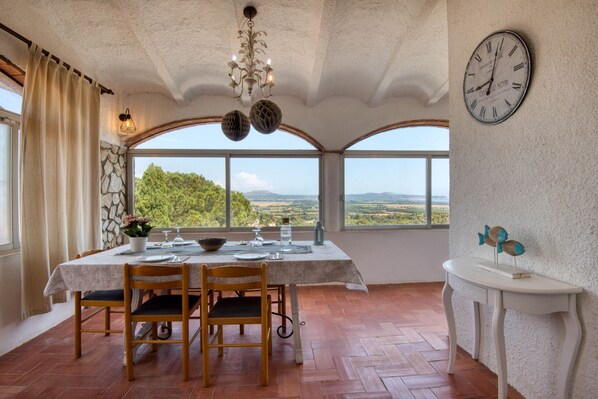 A dining area with a table set for two, a chandelier, a wall clock, and a view of the landscape through large windows.