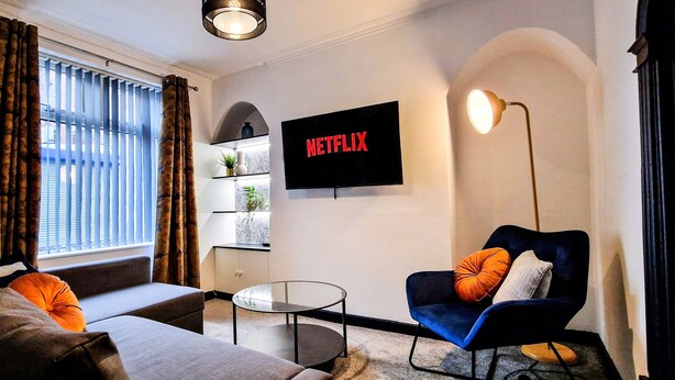 Ein modernes Wohnzimmer mit einer Couch, einem Sessel und einem Glastisch. Ein Flachbildfernseher an der Wand zeigt das Netflix-Logo.