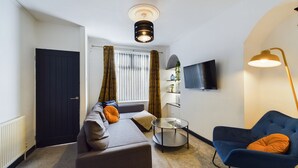 Ein modernes Wohnzimmer mit einer Couch, einem blauen Sessel, einem runden Couchtisch, einem an der Wand montierten Fernseher und einer Stehlampe.
