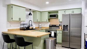 Uma cozinha compacta com armários verdes, uma geladeira de aço inoxidável, micro-ondas e pia. Há dois banquetas pretas e uma lixeira.