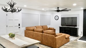 Uma sala de estar moderna com sofá seccional, mesa de jantar, lustre, ventilador de teto e televisão.