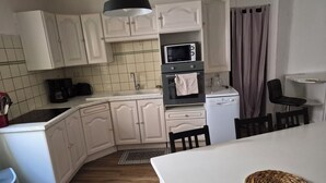 A compact kitchen with white cabinetry, a microwave, a dishwasher, and a dining area with a table and chairs. - Maison de Village (Puimisson)