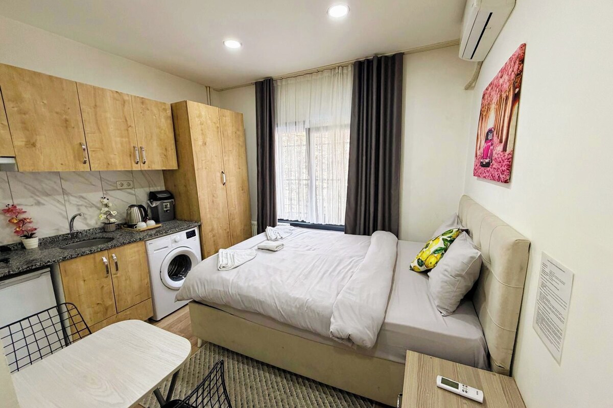 A compact hotel room with a bed, a small table, a washing machine, and a kitchenette.