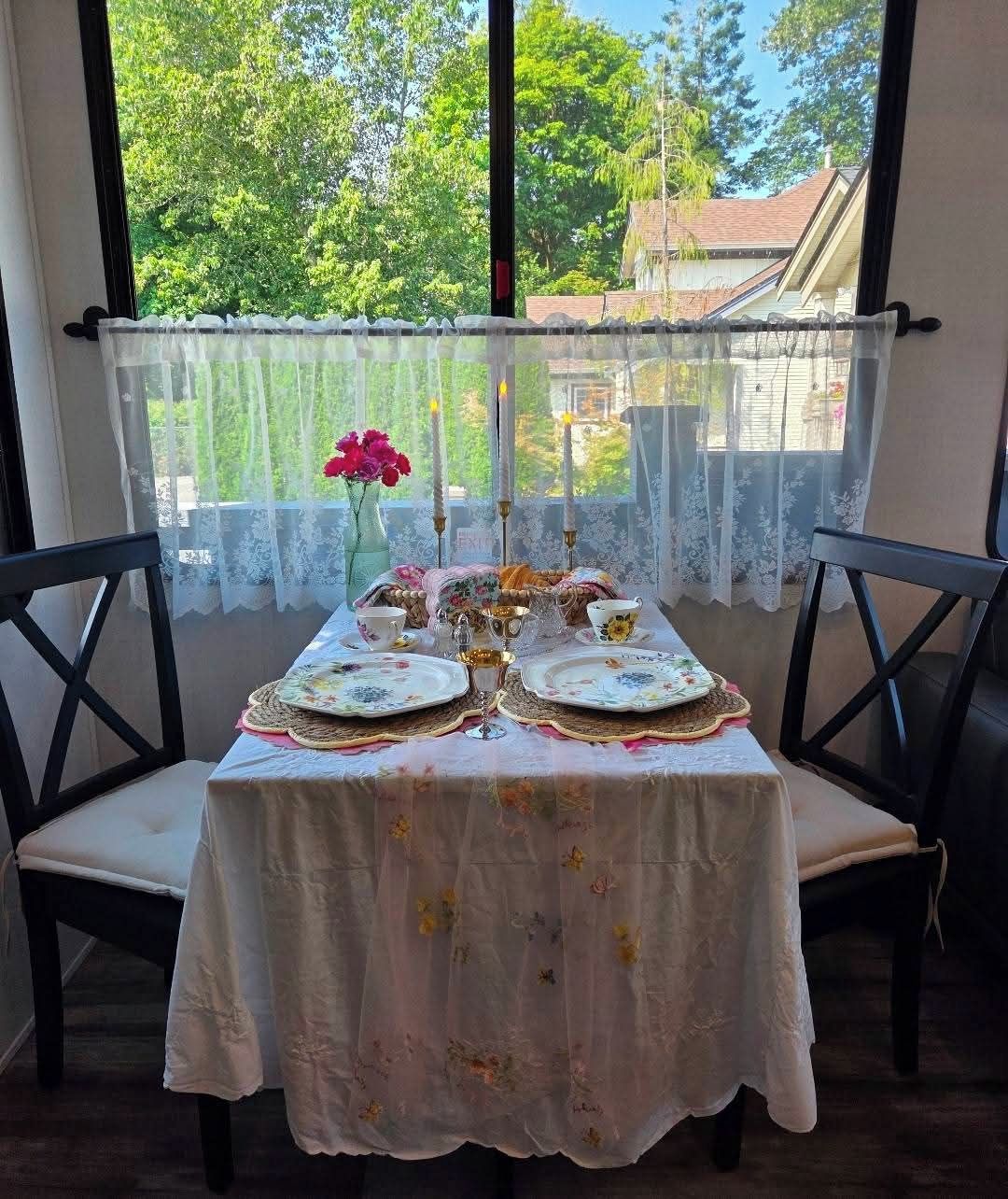 A table set for tea with floral tablecloth, lace curtains, and a vase of flowers.