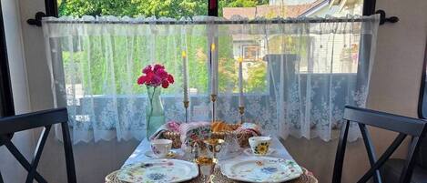 A table set for tea with floral tablecloth, lace curtains, and a vase of flowers.