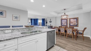 A modern kitchen with white cabinets, granite countertops, and a dishwasher. There is a dining area with wooden chairs and a table. - Ocean view 3 BR/3BA condo!  Royale Palms 12th floor! Perfect for families! (Myrtle Beach)