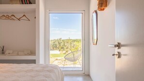 Une chambre avec un grand lit, une porte donnant sur un balcon équipé d’une chaise, et une vue sur un paysage verdoyant.