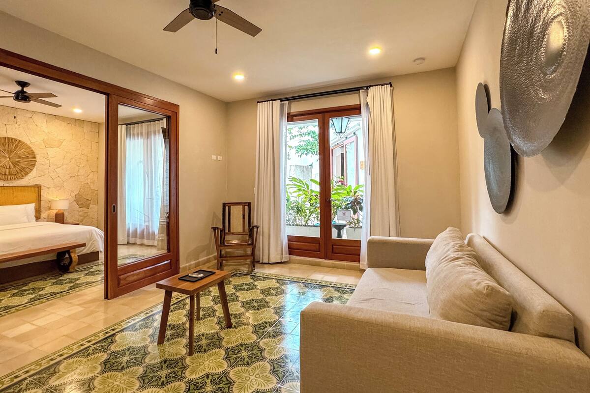 A hotel room with a bed, a sofa, a small table, a chair, a ceiling fan, and a view of the outside through a glass door.