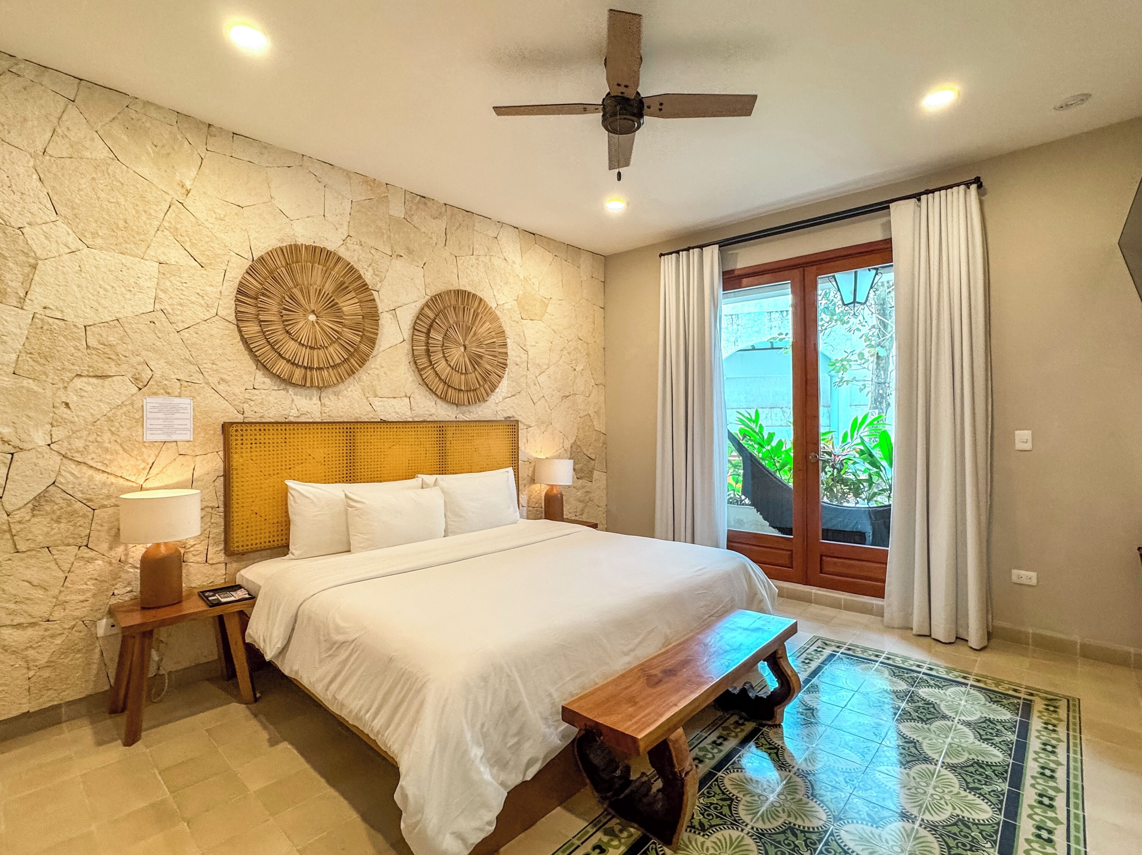 Un dormitorio con pared de piedra, dos adornos circulares en la pared, una cama con ropa blanca, un banco de madera, un ventilador de techo y vistas a la vegetación a través de una puerta de cristal.
