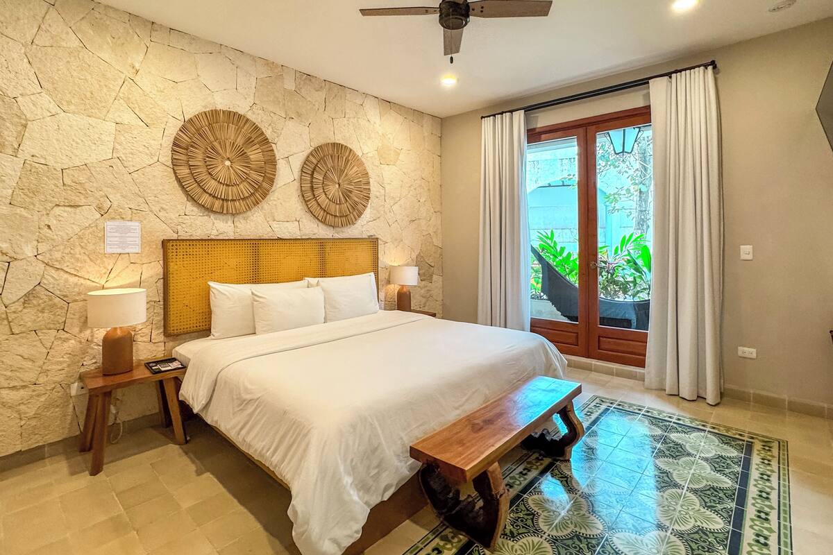 A bedroom with a stone wall, two circular wall decorations, a bed with white linens, a wooden bench, a ceiling fan, and a view of greenery through a glass door.