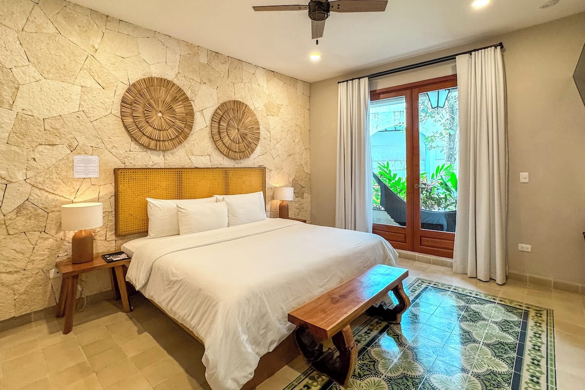 Un dormitorio con pared de piedra, dos adornos circulares en la pared, una cama con ropa blanca, un banco de madera, un ventilador de techo y vistas a la vegetación a través de una puerta de cristal.