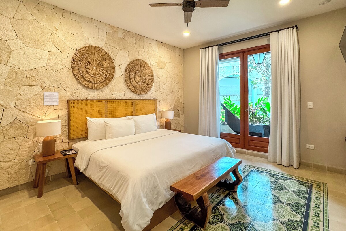 A bedroom with a stone wall, two circular wall decorations, a bed with white linens, a wooden bench, a ceiling fan, and a view of greenery through a glass door.