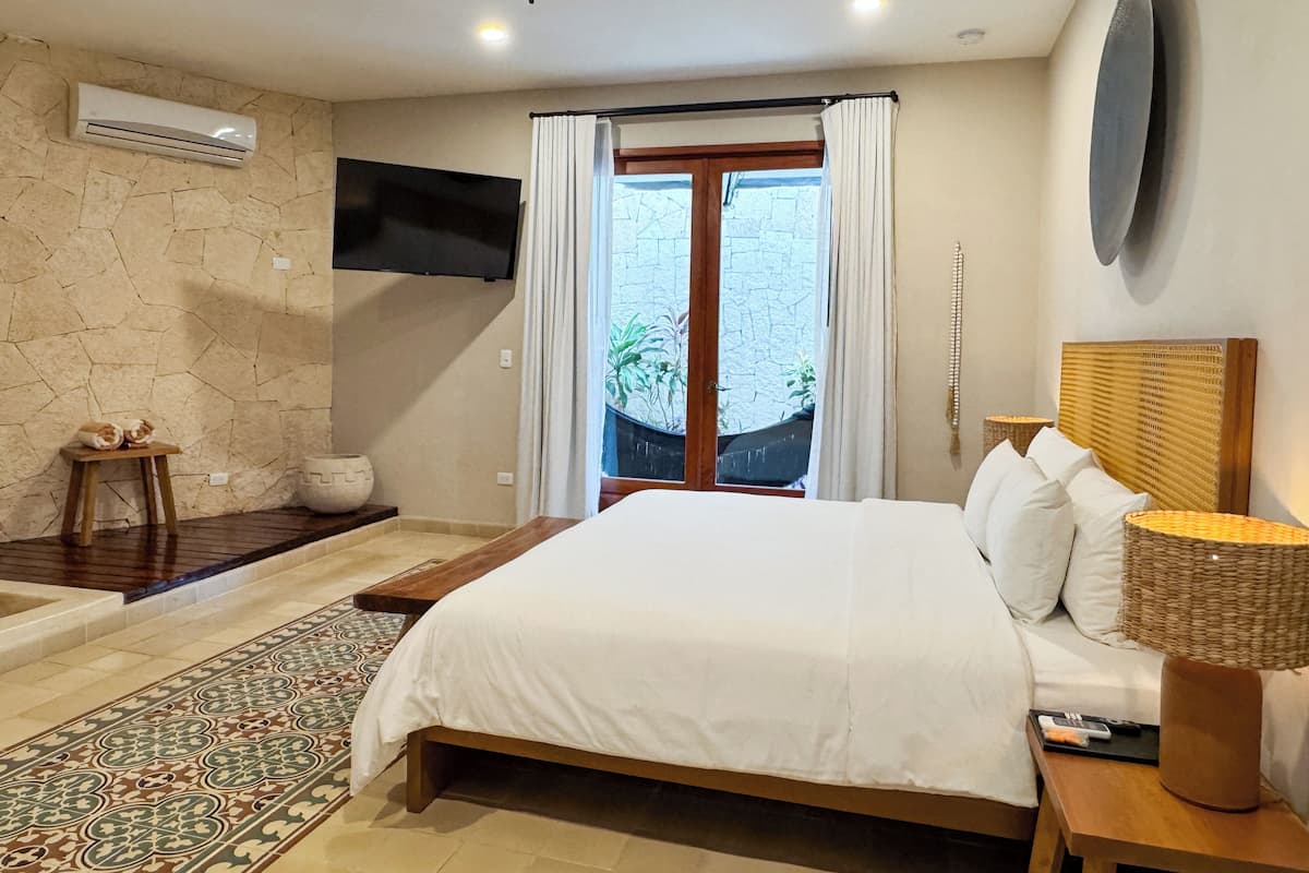 A bedroom with a bed, a TV mounted on the wall, a window with curtains, a stone wall, and a small table with a bowl.